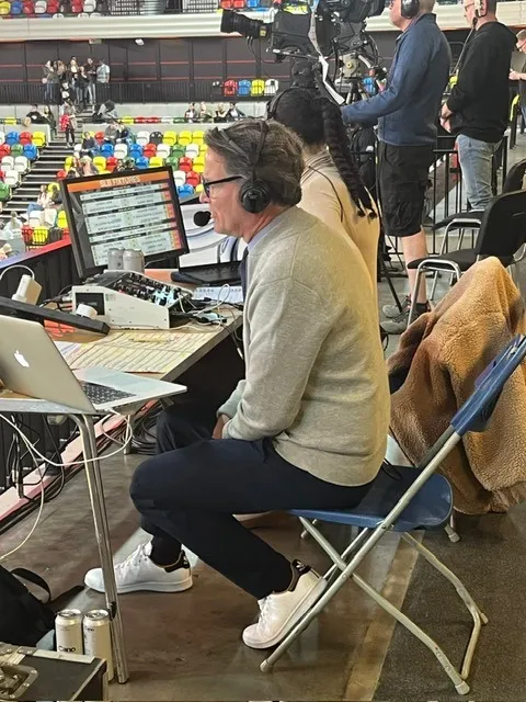 Todd Harris in a headset at an outside-broadcast desk, crowd and cameras behind him.
